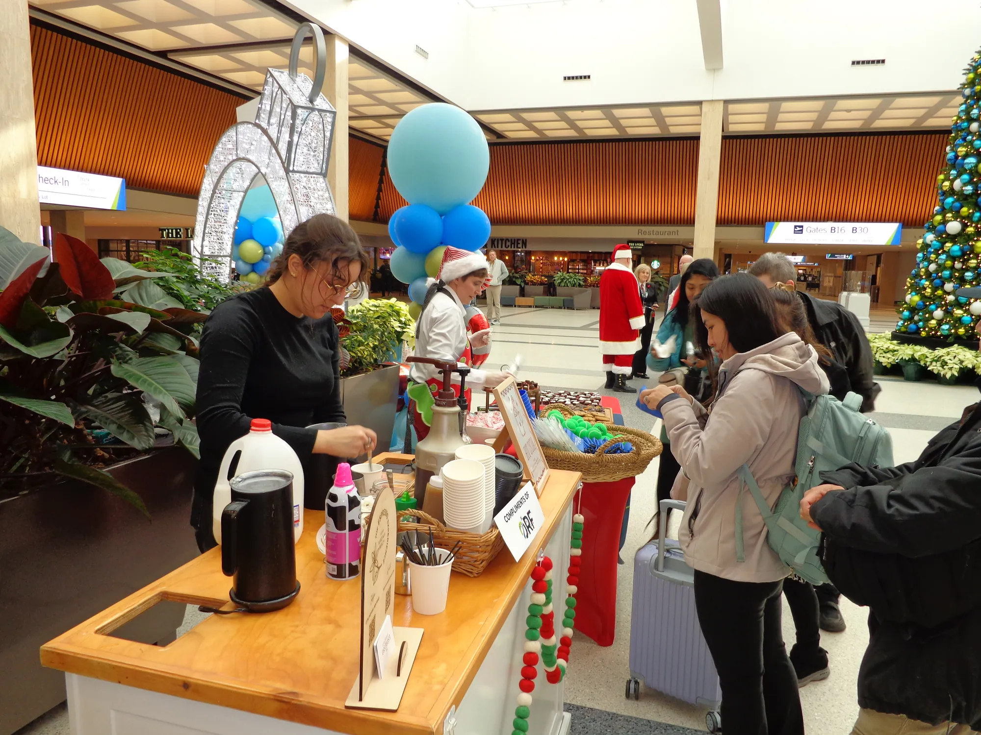 Barista serving travelers at a holiday hot chocolate and coffee station beside a Christmas tree