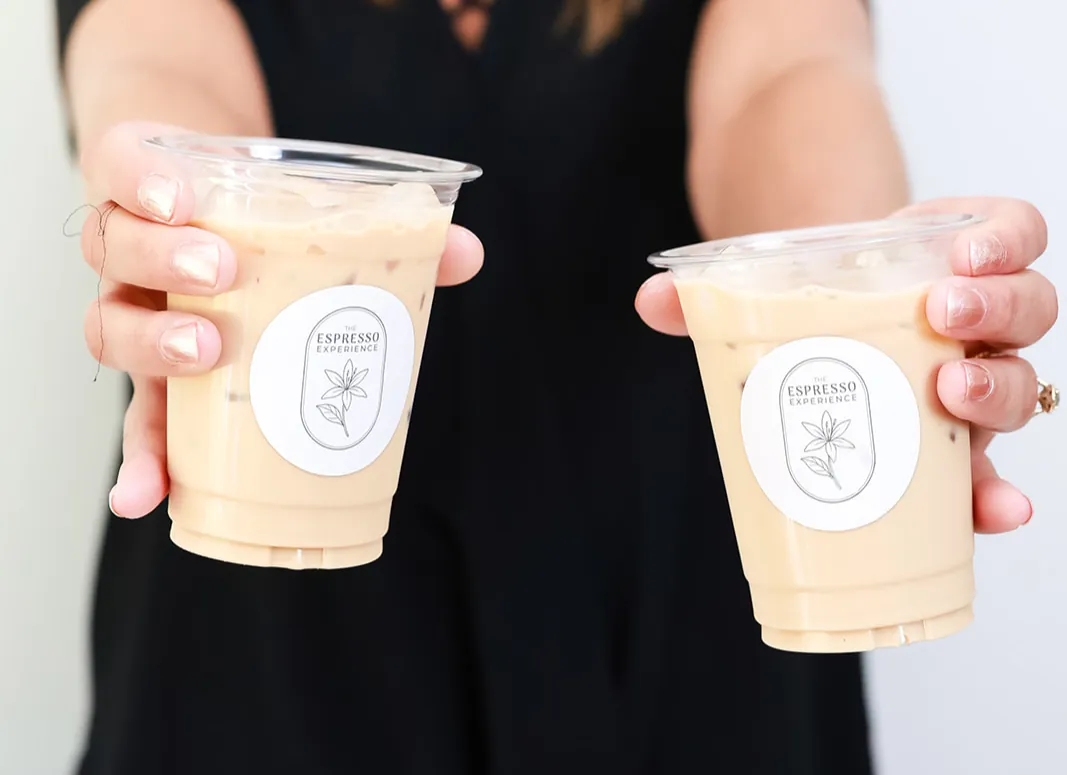 Two iced lattes in branded The Espresso Experience cups held out toward the camera