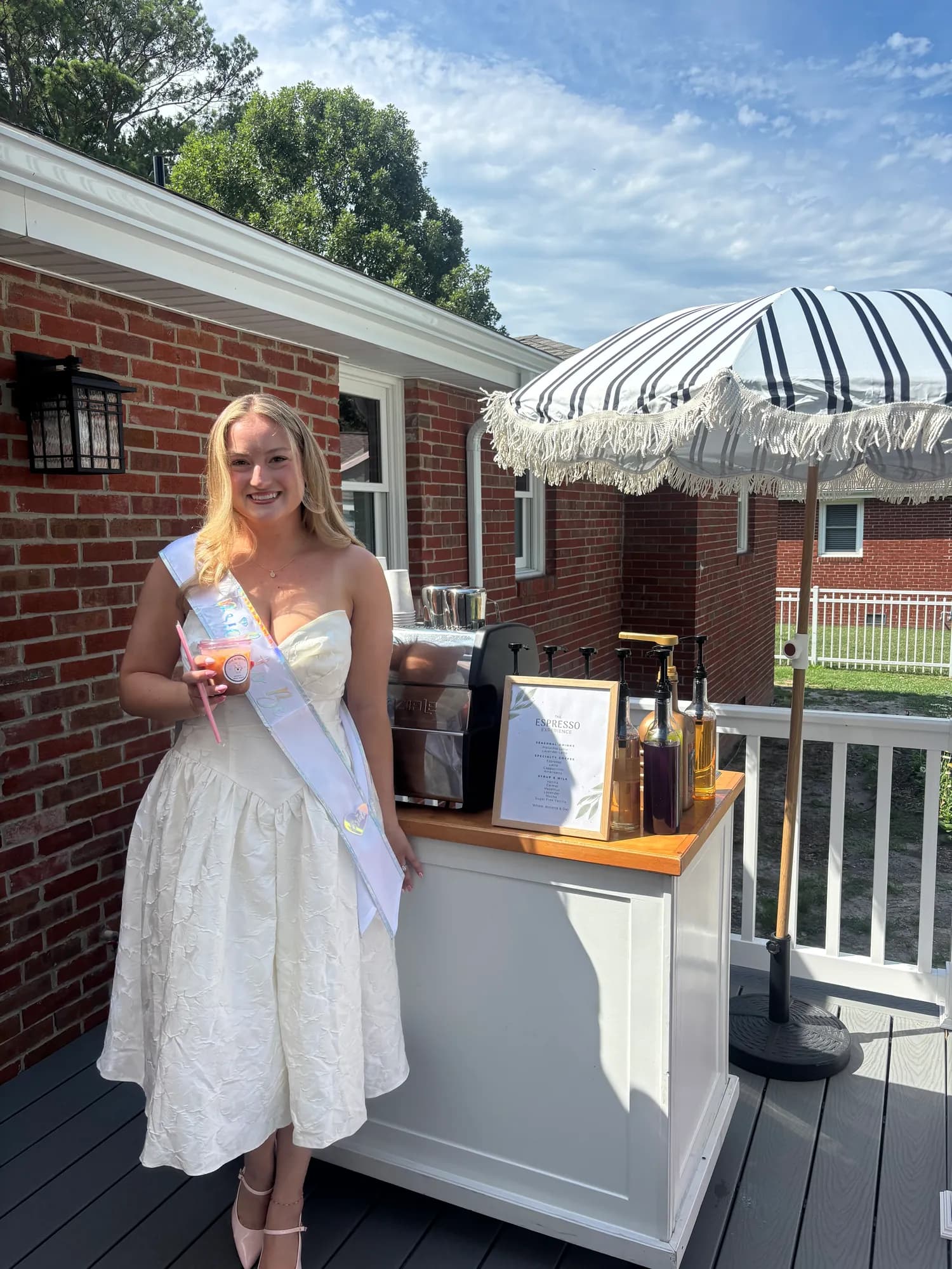 Guest of honor in a white dress and sash holding an iced drink beside the espresso cart